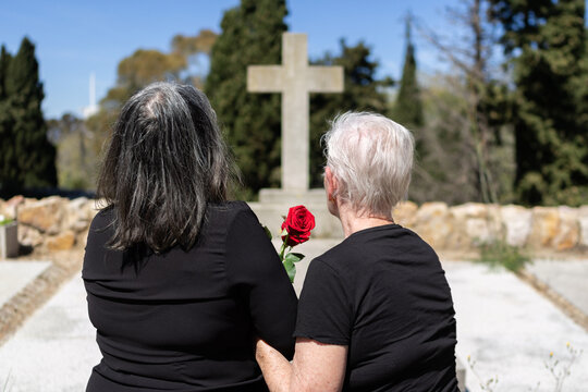 family women wearing black clothing and holding a red rose are in a burial funeral praying to a sacred christian stone cross in a cemetery
