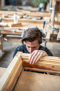 Craftsman finishing working on antique wooden frame restoration in workshop. Man woodworker sitting at workbench attentively looking checking fitting of wooden parts, assessing accuracy.