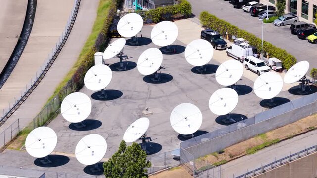 Satellite dish antenna farm seen from above. Aerial view of white parabolic antennas at a broadcast teleport ground station. Telecommunication technology and global data network on sunny day.