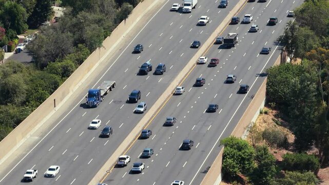 Aerial view of busy multi lane freeway with cars and semi trucks moving in both directions. Highway traffic flow between concrete sound walls and green trees under bright summer sunlight.