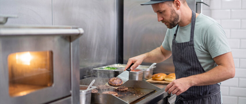 Chef flipping burger patty on griddle in a commercial stainless steel restaurant kitchen with steam rising from fresh beef, ideal for fast food marketing and menu promotion