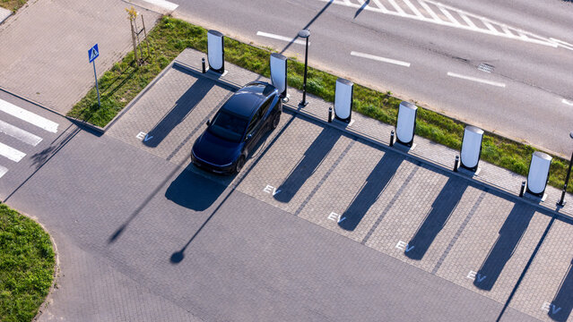 Aerial view of an electric vehicle charging station with a dark car parked at a charging point, featuring paved parking spaces with EV markings and a nearby road in Gdansk, Poland.