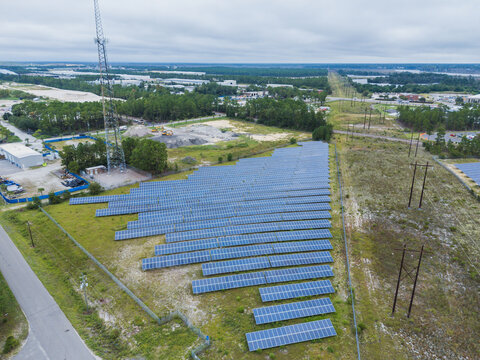 Aerial view of a solar farm with rows of photovoltaic panels installed on a grassy field, featuring a communication tower and surrounding trees in Wilmington, North Carolina, United States.