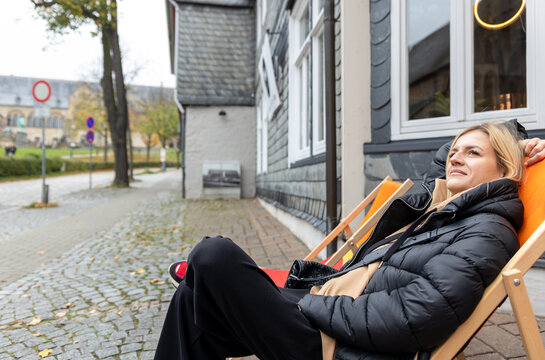 Relaxed woman sits on deck chair outside historic building in Goslar, Germany, enjoying quiet travel moment. Candid tourist lifestyle scene captures leisure, autumn mood and local urban charm