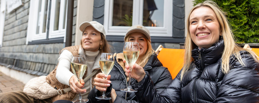 Three smiling women toast with sparkling wine outside cafe in Europe, enjoying friendship, travel and celebration. Candid lifestyle scene captures leisure, reunion, happiness and autumn atmosphere.