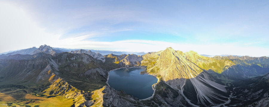 Aerial view of the turquoise waters of Lunersee surrounded by rugged alpine peaks, steep rocky slopes, and lush mountain meadows in Vandans, Vorarlberg, Austria.