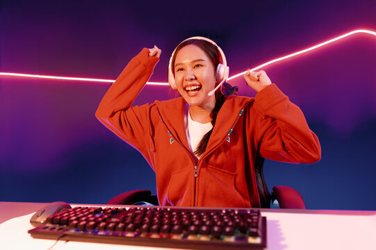 Host channel of smiling beautiful Asian girl streamer playing with raising fist up winning on Esport skilled team players wearing headphones pastel color, fighting online game at neon room. Stratagem.