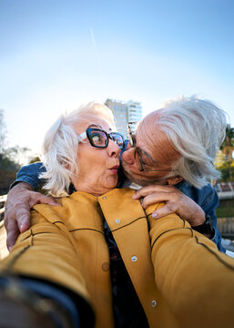 Playful senior couple taking a close selfie as the man kisses the surprised woman on the cheek. Wide-angle urban scene conveying affection, humor, companionship and active aging.