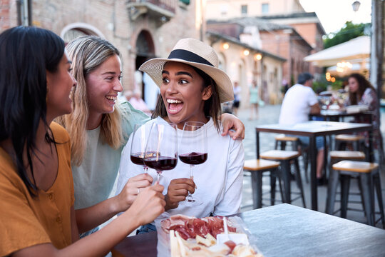 Three happy multi-ethnic women friends are toasting red wine glasses while sitting at a table of an outdoor bar in a european city, eating a plate of cold cuts and cheese