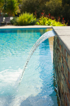 Elegant swimming pool with a cascading water feature flowing from a stone wall into clear turquoise water, surrounded by lush Mediterranean garden vegetation. Bright sunny outdoor scene with a relaxin