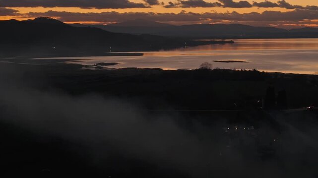 Aerial view of Lago Trasimeno at sunset with mist over the water and rolling hills in the Province of Perugia, Umbria, Italy.