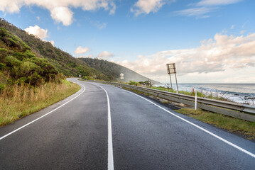 Obraz premium Empty winding coast road under blue sky with clouds at sunset in autumn. A thin mist is drifting from the ocean.