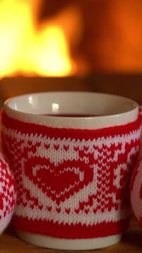 Mugs in knitted woolen covers with a Christmas ornament are placed in front of the fireplace. Xmas decorations on a wooden table in front of a flame burning in the fireplace. Vertical video