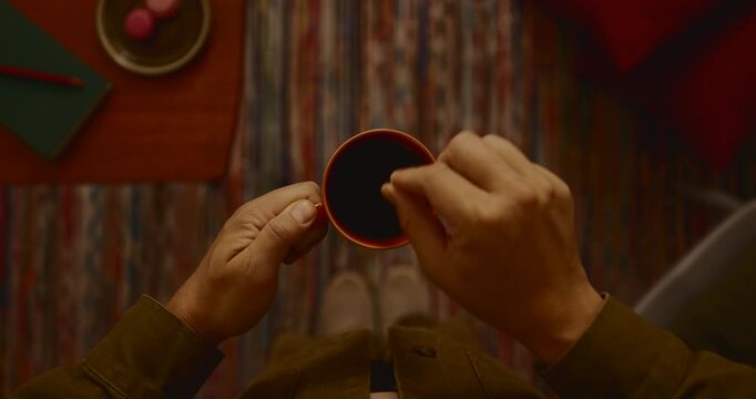 Top view of hands holding a coffee cup and stirring tea with a spoon above a colorful carpet background, person enjoying warm beverage at home, cozy indoor lighting, relaxed morning lifestyle shot.
