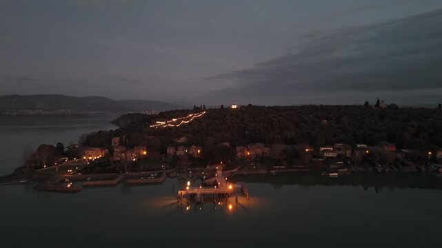 Aerial view of the illuminated pier and historic village of Isola Maggiore on Lake Trasimeno at dusk in Tuoro sul Trasimeno, Umbria, Italy.