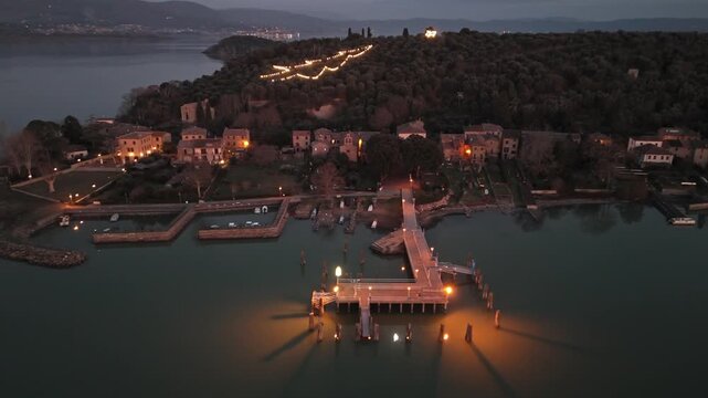 Aerial view of Isola Maggiore on Lake Trasimeno at dusk with an illuminated pier and village lights in Tuoro sul Trasimeno, Umbria, Italy.