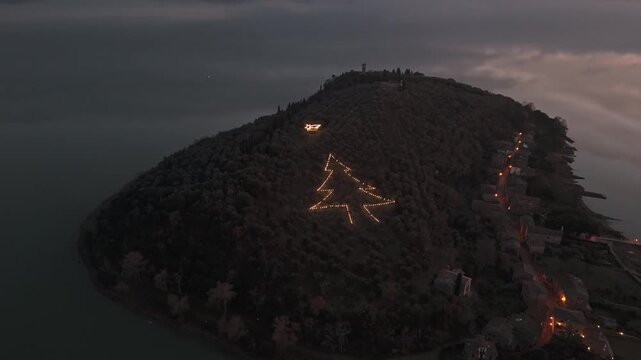 Aerial view of Isola Maggiore on Lake Trasimeno featuring a large illuminated Christmas tree shape on the hillside at dusk in Tuoro sul Trasimeno, Umbria, Italy.