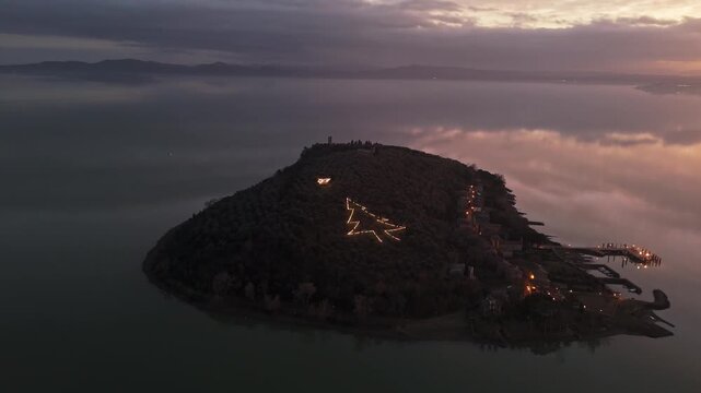 Aerial view of the illuminated tree light installation on Isola Maggiore at dusk, surrounded by the calm waters of Lake Trasimeno in Tuoro sul Trasimeno, Umbria, Italy.
