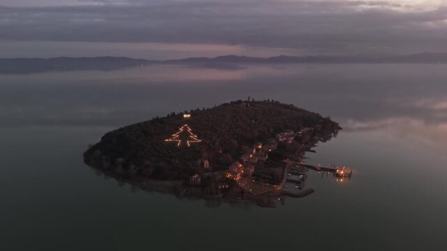 Aerial view of Isola Maggiore on Lake Trasimeno featuring a large illuminated Christmas tree shape on the hillside at dusk in Tuoro sul Trasimeno, Umbria, Italy.