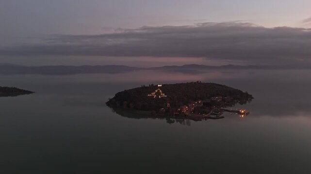 Aerial view of Isola Maggiore on Lake Trasimeno featuring a large illuminated Christmas tree light installation at dusk in Tuoro sul Trasimeno, Umbria, Italy.