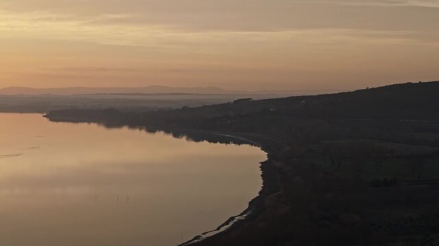 Aerial view of the calm waters of Lake Trasimeno at sunset with Isola Maggiore in the distance and rolling hills along the shoreline in Tuoro sul Trasimeno, Umbria, Italy.