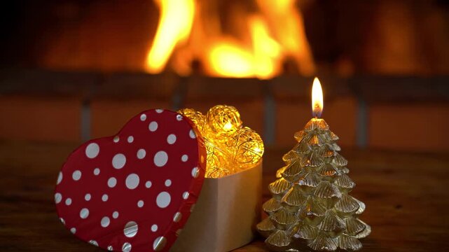 Candle and Christmas tree decorations are placed on a wooden table in front of a flame burning in the fireplace