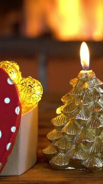 Candle and Christmas tree decorations are placed on a wooden table in front of a flame burning in the fireplace. Vertical video
