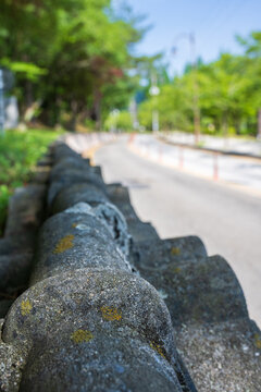 Close-up of weathered roof tiles on roadside stone wall extending along curved asphalt street in rural South Korea.