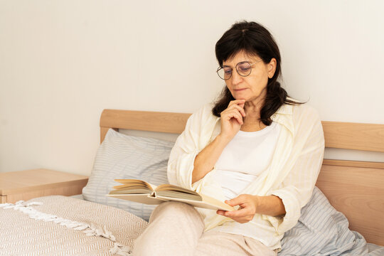 Mature adult woman wearing glasses relaxes in bed with book, enjoying her leisure time and an intellectual pursuit. Slow living, mindful consumption, analog hobby