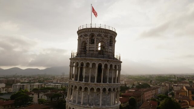 Aerial view of the iconic Leaning Tower of Pisa with its marble arches and a flag on top, set against the soft light of the golden hour over the historic city of Pisa, Toscana, Italy.