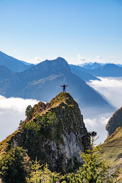 Aerial view of a person standing with arms outstretched on a rocky mountain peak in the Swiss Alps, surrounded by clouds and mountain ranges in Airolo, Ticino, Switzerland.