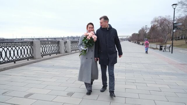 Romantic couple walking along riverside promenade while woman carries bouquet of pink roses