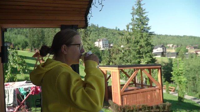 woman yellow hoodie glasses drinking coffee chatting terrace mountain cottage, enjoying panoramic view mountains nature. background, wooden gazebo tall green trees. warm summer day.