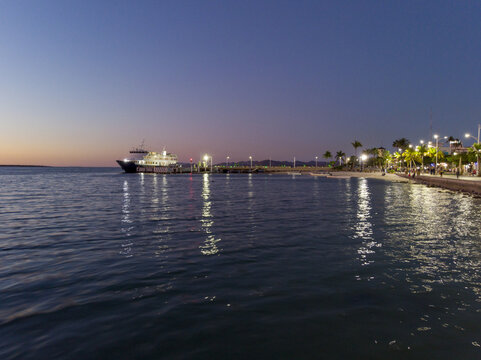 Aerial view of Malecon de La Paz at dusk with a ferry docked at the pier, illuminated palm trees, and street lights reflecting on the calm water in La Paz, Baja California Sur, Mexico.