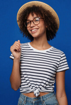 Smiling young African American woman with curly afro hair wears straw hat, glasses. Happy black female person poses in striped t-shirt, denim jeans against blue background. Joyful girl shows teeth,