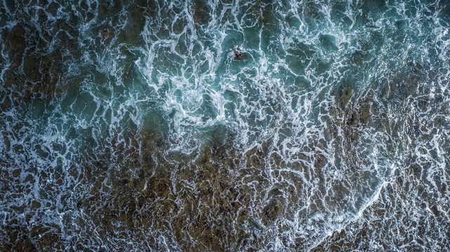 Aerial view of ocean waves crashing over a rocky reef shoreline with turquoise water and white sea foam in Bajawa, Nusa Tenggara Timur, Indonesia.