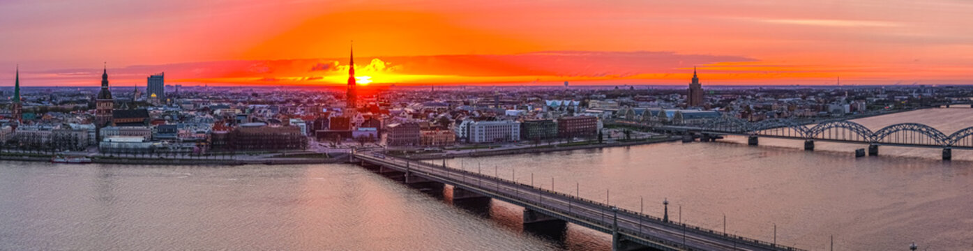 Aerial view of the Riga skyline at sunset, featuring the Daugava River, the Stone Bridge, and historic church spires under a vibrant orange sky in Riga, Latvia.