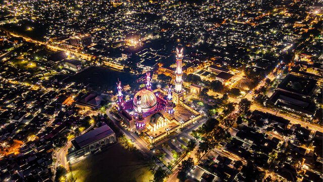 Aerial view of the illuminated Islamic Center NTB - Hubbul Wathan Great Mosque at night, surrounded by the glowing city lights of Kecamatan Selaparang, Nusa Tenggara Barat, Indonesia.