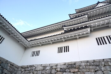 白い漆喰の壁と黒い瓦屋根が特徴的な日本の城郭建築をローアングルから捉えた一枚。青空の下、重厚な石垣の上に築かれた美しい建物のディテールが際立つ。 © BRUCE