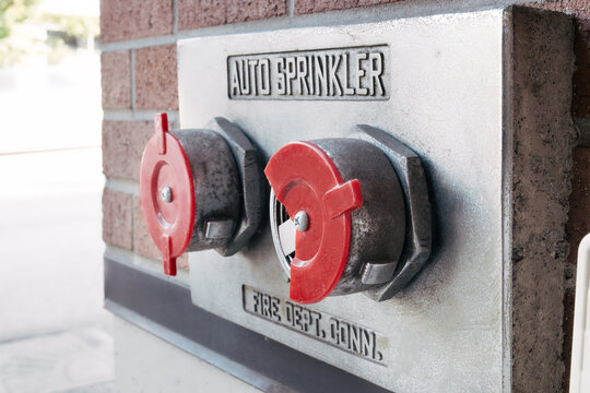 Auto sprinkler standpipe fire department connection with broken cap. Exposed connector vulnerable to debris and tampering. Fire safety compliance concept on brick wall exterior. Selective focus.