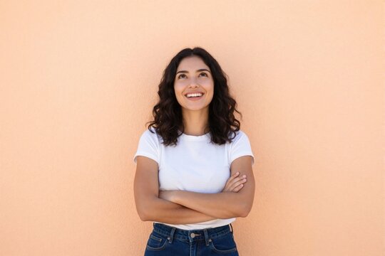 Young latin woman stands against plain beige wall smiling up with crossed arms. She looks hopeful and happy thinking about future goals or success.