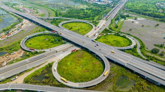 Aerial view of the JNPT Interchange featuring complex highway ramps and heavy truck traffic surrounded by lush green wetlands in Navi Mumbai, Maharashtra, India.