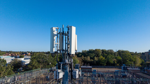 Aerial view of a telecommunications phone mast with antennas and equipment, set against a clear blue sky above a power substation in Camberley, England, United Kingdom.