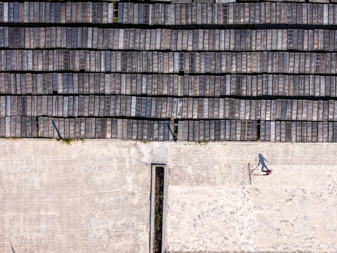 Aerial view of a worker raking crops on a large drying ground with rows of drying noodles on racks in Polanharjo, Central Java, Indonesia.