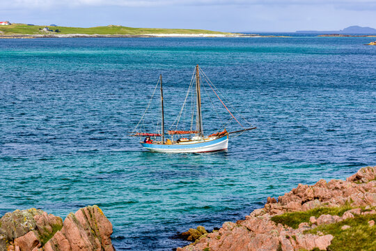 Small two-masted sailing boat floating on clear blue ocean water, pink granite rocks in foreground and grassy coast in distance