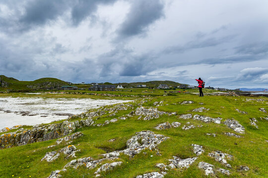 Hiker with backpack photographing coastal scenery on remote island, wide view of rocky outcrops, sandy beach and distant houses under dramatic clouds