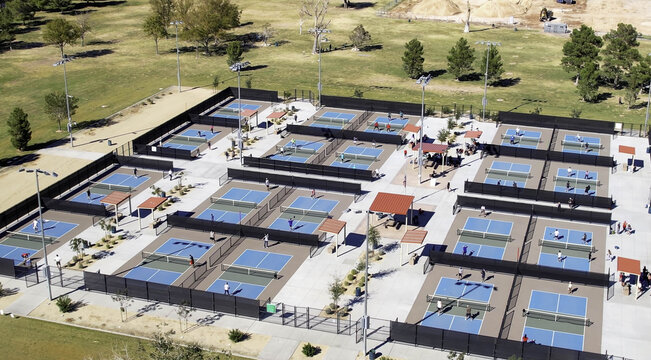 Aerial view of several rows of pickleball courts with players on the blue and green surfaces, surrounded by fencing and park grounds in Valencia, California, United States.
