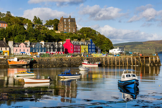 Brightly painted houses lining the harbor of a traditional Scottish fishing village, reflections of boats shimmer on the still sea water surface