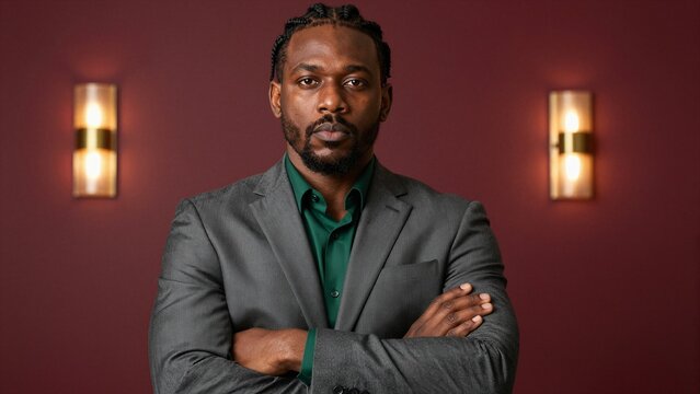 A serious executive man with braided hair and beard stands confidently with arms crossed against a deep red wall illuminated by warm sconces.