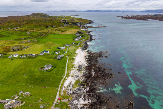 Aerial view of coastal village and sandy beach on Isle of Iona, Scotland, showing turquoise ocean waters along a rocky shore with green farmland